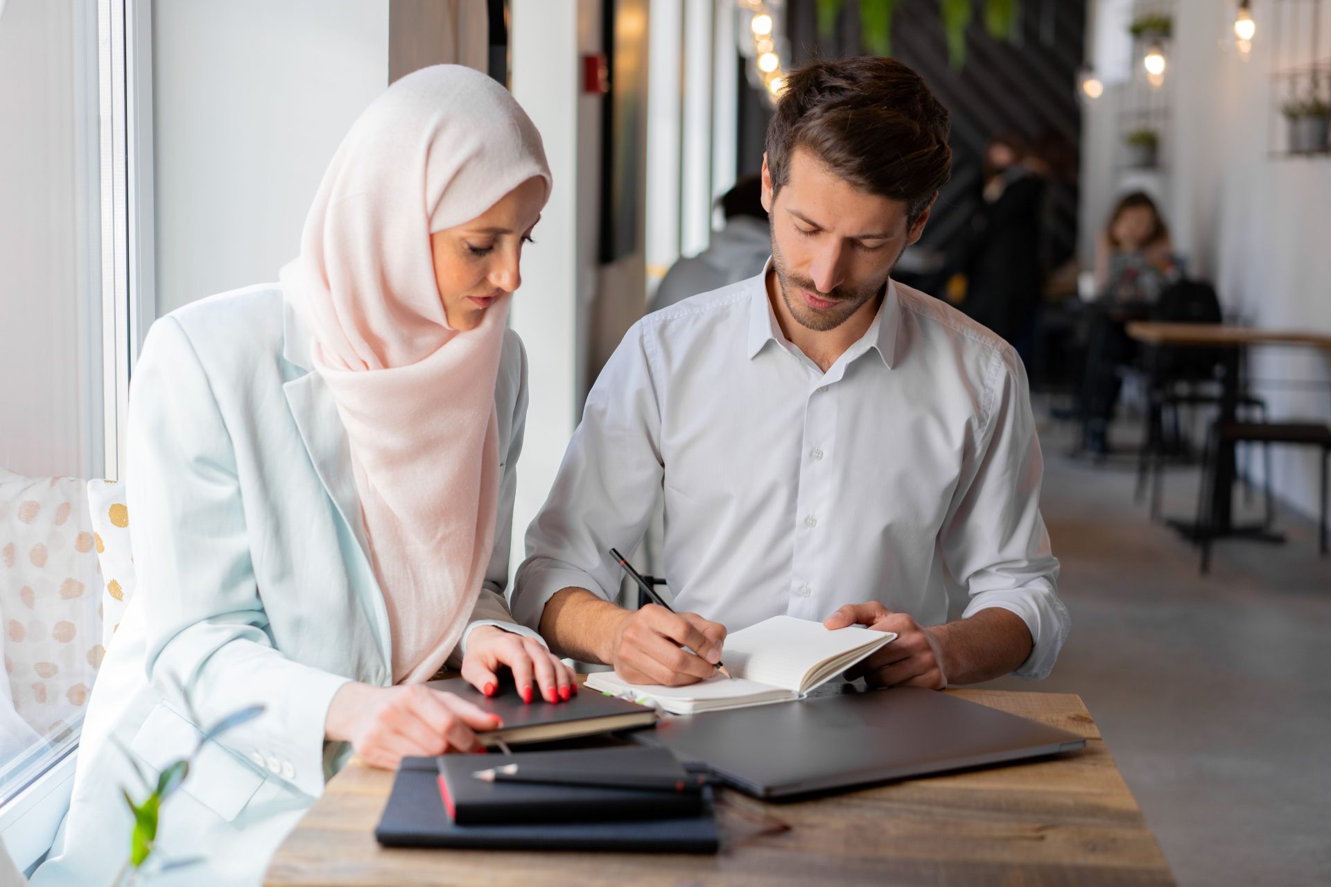 A man and woman in business attire collaborate at a table indoors, with a focus on writing notes.
